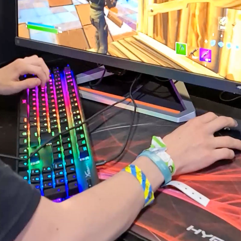 Hands on multi coloured keyboard and mouse besides a gaming PC screen