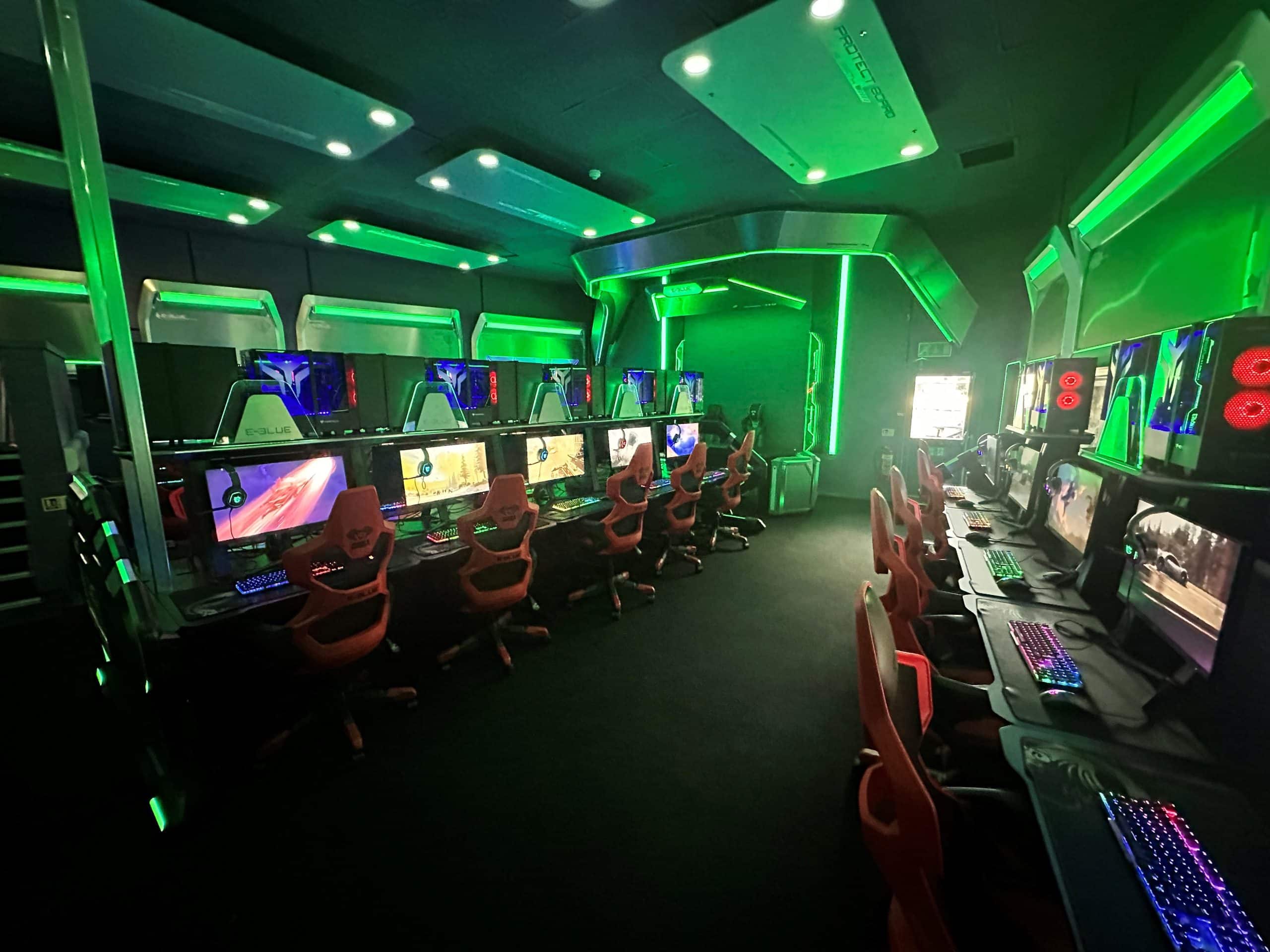 8 gaming stations with gaming chairs, screens, computers, keyboards and headphones in esports arena. Green lights along ceiling